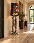 Sculptural floor lamp with two antique silver-tone figures and a dark floral shade, standing on a stone floor in a spacious hallway.